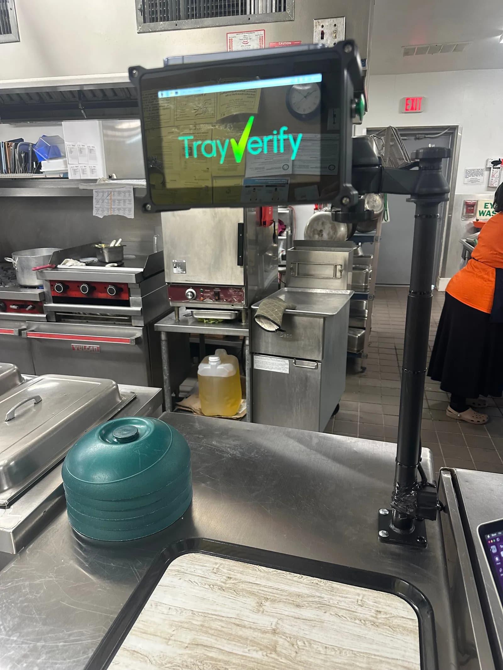 TrayVerify device on a black pole mount at the end of a stainless tray-line table, kitchen ranges and warming cabinets behind