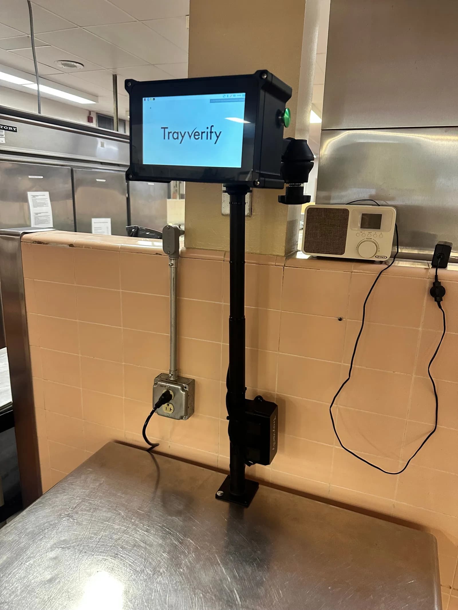 TrayVerify device on a black pole rising from a stainless counter in front of a tiled wall